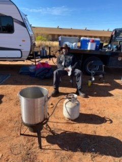 Person sitting by a camping setup with a propane burner and a large pot. A trailer and flatbed truck are in the background.