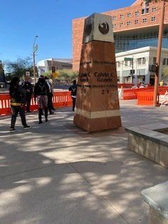 Monument with a phoenix emblem, people standing nearby. Outdoor setting with construction barriers.