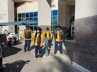 Group of people in yellow vests, blue pants, and masks posing outside a building with motorcycles.