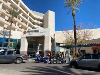 Hospital entrance with sign, palm trees, and vehicles parked out front. Sunny day.
