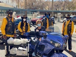 Motorcycle club members deliver food on a blue motorcycle to a woman in a parking lot. Riders wear vests and cowboy hats.