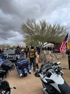 Motorcyclists wearing vests at an outdoor gathering with a US flag. Bikes parked in front. Cloudy sky.