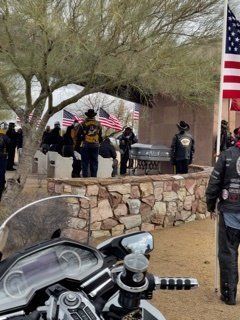 Motorcycle riders at a memorial service with a casket, US flags, and stone wall in an outdoor setting.