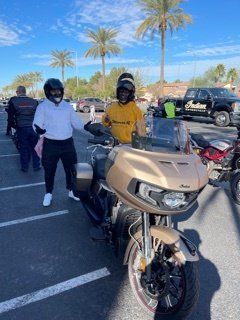 Two people wearing helmets pose next to a gold motorcycle in a sunny parking lot.