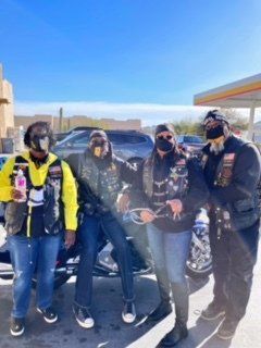 Four people in motorcycle gear stand outdoors at a gas station, posing for a photo.