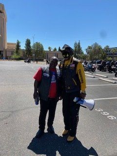 Two men standing outside, one holding a megaphone. They are near motorcycles on a sunny day.