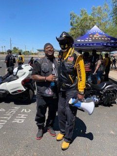 Two men pose outdoors near motorcycles; one holds a megaphone. They wear vests, jackets, and hats under a blue canopy.