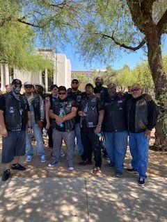 Group of people, wearing vests, standing outside. Some have arms around each other. Trees and a building in the background.