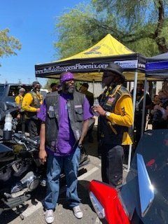 Two men in vests and hats stand near a black motorcycle and a yellow tent at an outdoor event.