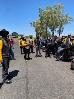Group of motorcyclists in yellow vests gathered outdoors near a building under a bright sky.
