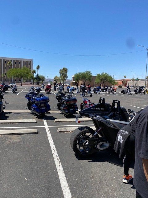 Motorcycles parked in a lot under a clear blue sky. People standing nearby.