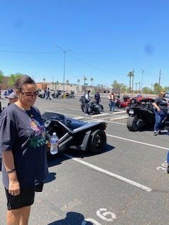 Woman standing near a black Slingshot; other motorcycles and people in a parking lot. Blue sky overhead.