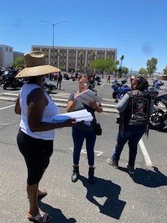 Three people review papers in a parking lot with motorcycles. Sunny day, building in background.