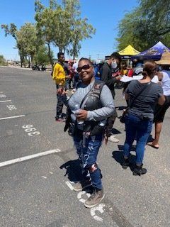 Woman in a biker vest and jeans smiles, holding a drink, outdoors among other people.