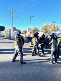 Motorcyclists in dark leather gear stand outside, some wearing helmets and face coverings.