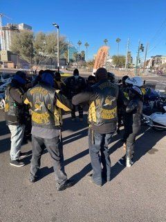 Group of bikers in black vests with gold lettering, gathered in a circle on a city street.
