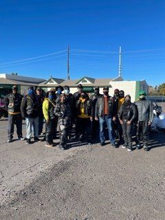 Group of people, many in leather jackets, standing outside on a sunny day.