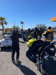 Motorcycle riders in black and yellow vests standing near motorcycles in front of a gas station under a blue sky.