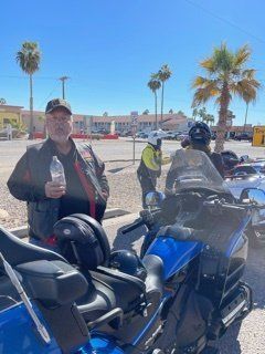 Man holding water bottle stands next to a blue motorcycle. Others wear helmets near palm trees and buildings.