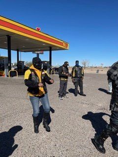 People in black and yellow vests with masks stand near a gas station. Clear, sunny day.