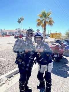 Two people pose outdoors near parked motorcycles. One wears biker gear, the other a plaid shirt. Blue sky, palm trees.