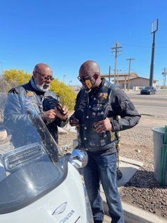 Two men in biker vests inspecting something near a parked motorcycle on a sunny day.