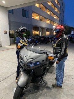 Two people in motorcycle helmets stand near a silver motorcycle in front of a building at dusk.