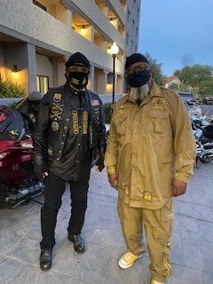 Two people in motorcycle club attire pose outside a building, wearing face masks. One in black jacket, one in tan suit.