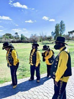 Group of bikers in yellow and black attire, wearing hats and face coverings, outdoors in a grassy area under a blue sky.