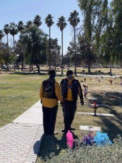 Two people in yellow jackets walk in a cemetery, alongside a stone path and flower arrangements.