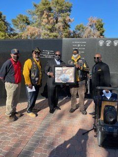 Group of men holding a framed item in front of a memorial wall.