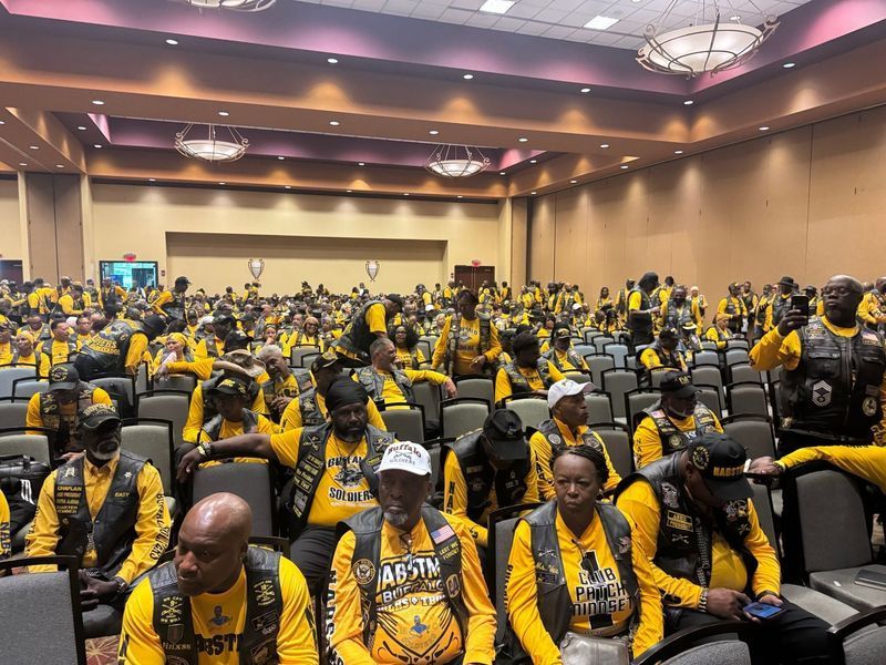 Large group of people in a conference room, many wearing yellow and black vests, seated, listening.