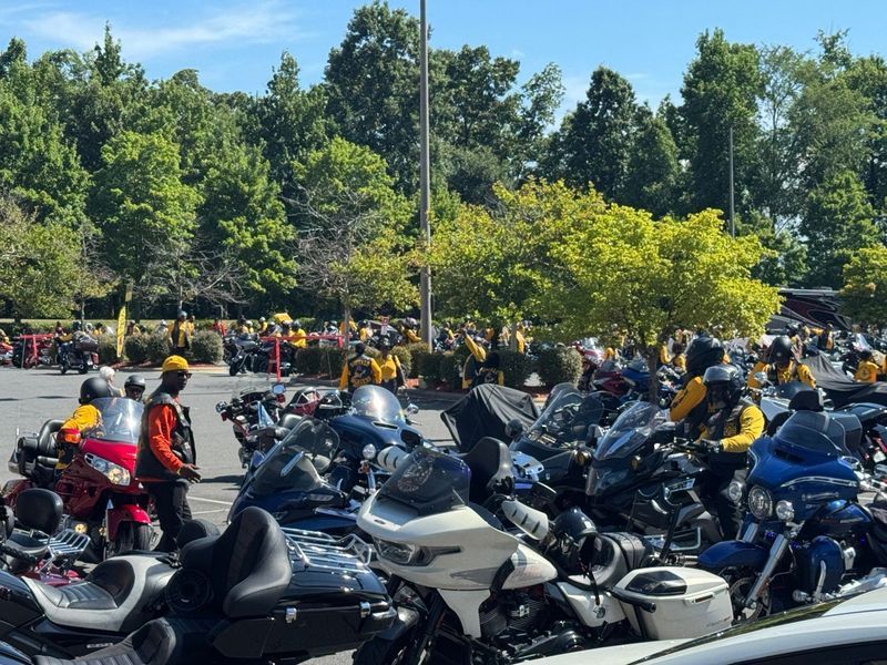 Motorcyclists in yellow and black gear gather in a parking lot on a sunny day.