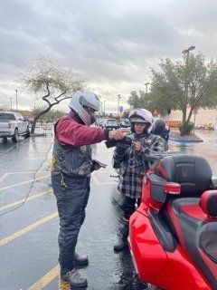 Two people near a red motorcycle. One is pointing to the other, both in helmets, standing in a parking lot on a rainy day.