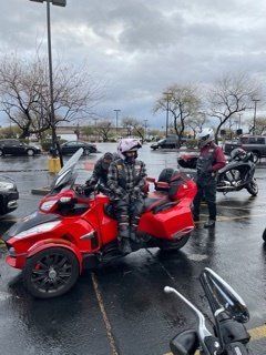 A red three-wheeled motorcycle in a parking lot with two riders in protective gear on a cloudy day.