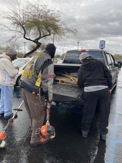 Three people loading equipment, including a weed wacker, into the back of a black pickup truck in a parking lot.