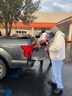 Two people loading items into a truck bed outside a Home Depot. Grey truck, rainy day.