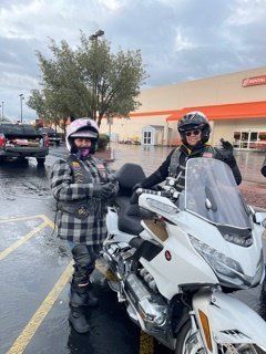 Two people in helmets and motorcycle gear pose next to a white motorcycle in a parking lot.