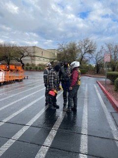 People in work attire stand in a parking lot. Cloudy sky. Wet pavement. Orange equipment visible.