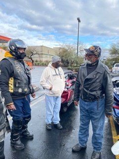 Three people conversing near parked motorcycles in a parking lot. Cloudy sky, pavement is wet. One wears helmet, vest.