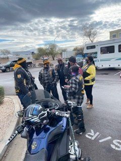 Group of people near a motorcycle; some wearing vests, talking outdoors in front of a RV.