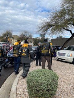 Group of people in black and gold vests and jackets, near motorcycles and a white car, outdoors.