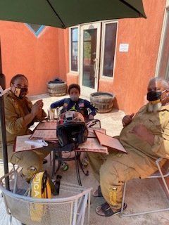 Three people seated at outdoor table with menus, wearing masks. Orange building background.