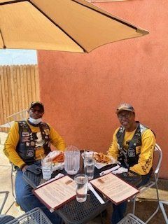 Two people in yellow shirts and vests sit at an outdoor table under an umbrella.