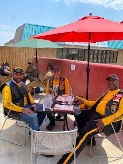 Three people in yellow shirts and vests sit at a table under a red umbrella, outdoor setting.