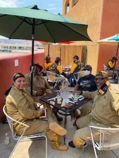 Group of people in tan outfits, seated at outdoor tables under umbrellas, conversing and socializing.