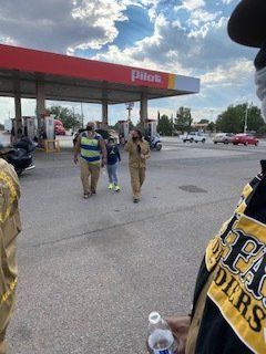 People walking near a Pilot gas station. One person holds a camera, others wear masks.
