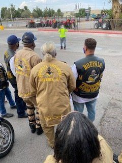 Motorcycle club members in tan outfits and vests stand in a line. A person in a neon green vest is facing them.