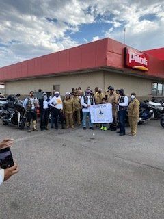 Group of people standing in front of a Pilot gas station, some wearing vests and holding a sign. Motorcycles are nearby.