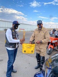 Two men with painted faces, in a parking lot, one receiving a yellow bag and wooden object.